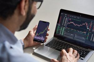 An image featuring a crypto broker using his laptop and phone watching the financial market