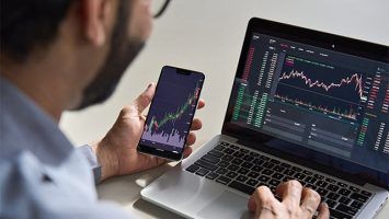 An image featuring a crypto broker using his laptop and phone watching the financial market